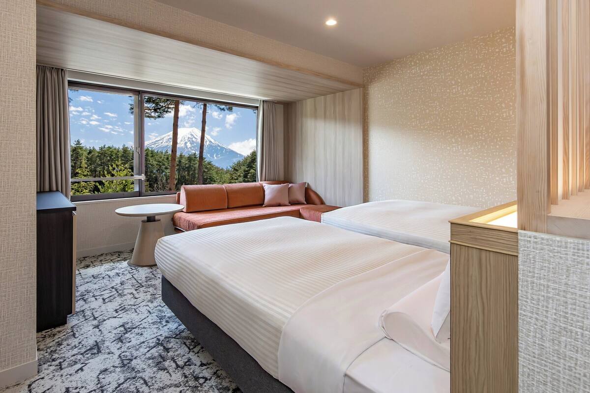 A hotel room with two beds, a small table, and a window with a view of a mountain.