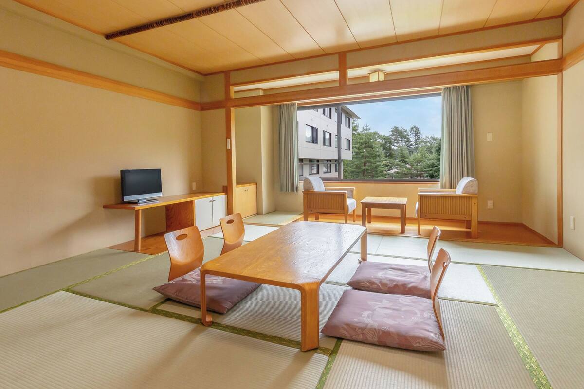 A traditional Japanese room with tatami flooring, a low table, chairs, a television, and a view of a building and trees through large windows.