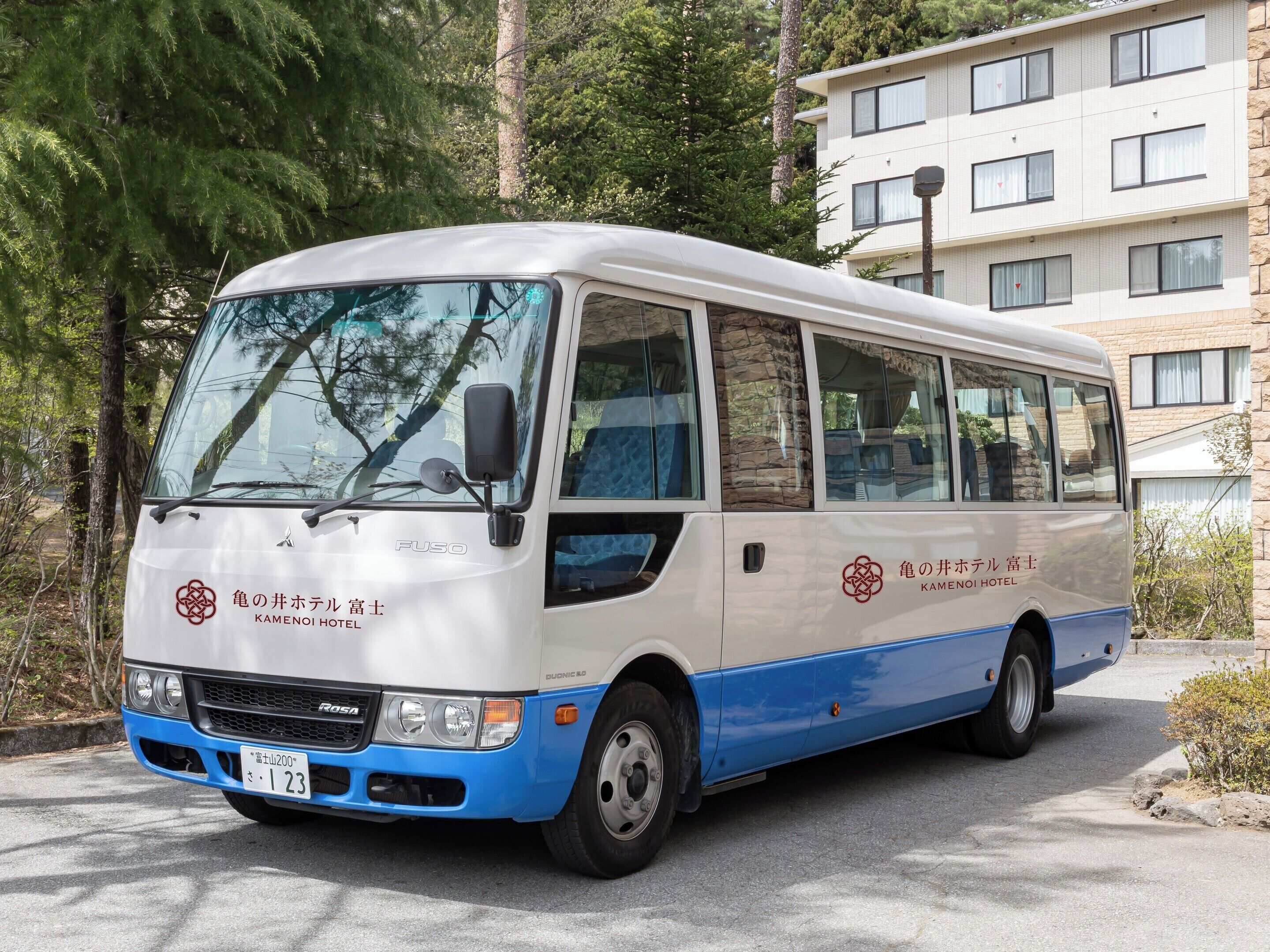 A white and blue bus with 'KAMENOI HOTEL' branding, parked on a street with trees and a building in the background.