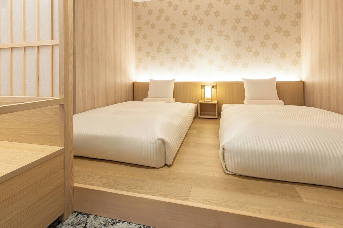 A hotel room with two beds, a nightstand with a lamp, wooden flooring, and a star-patterned wall.