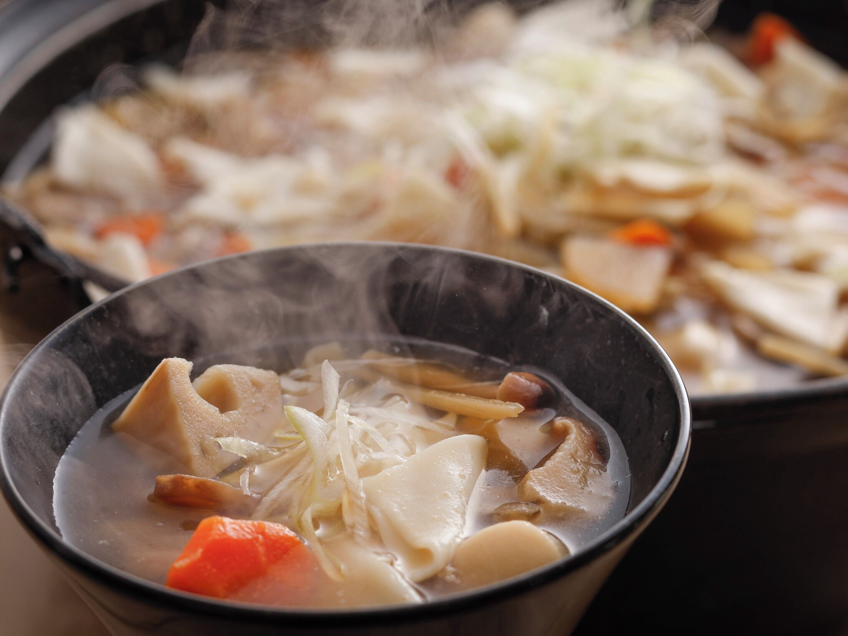 Un bol de sopa amb diversos ingredients, vapor que s'eleva del bol, i una paella amb més menjar al fons.