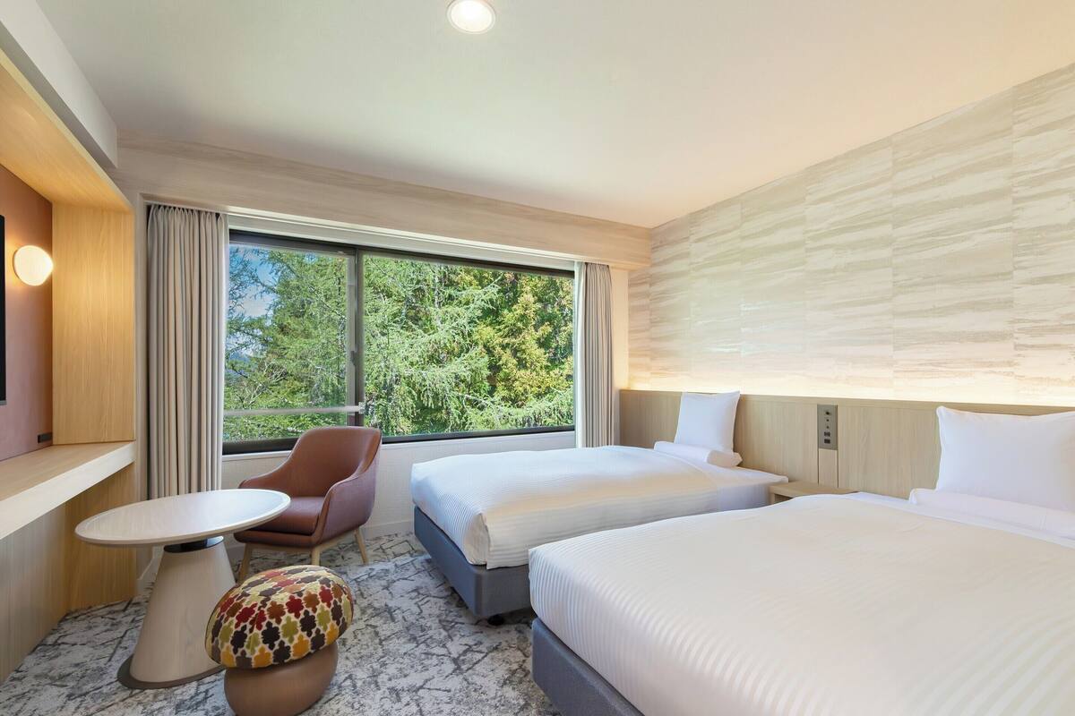 A hotel room with two beds, a small table, a chair, and a window with a view of trees.