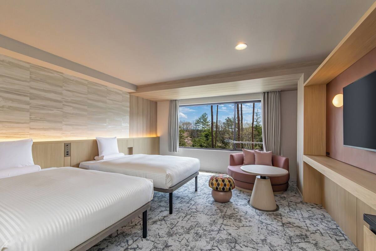 A hotel room with two beds, a sofa, a small table, a TV, and a window with a view of trees.