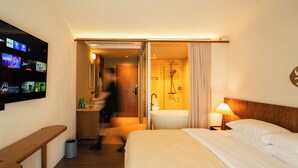 Habitación de hotel con una cama grande, un banco de madera, un televisor montado en la pared y un baño con ducha y bañera.