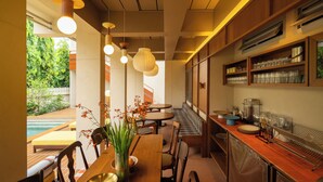 Un comedor moderno con una mesa de madera, sillas y lámparas colgantes. Se ve una piscina a través de la ventana, y un área de descanso con un banco.
