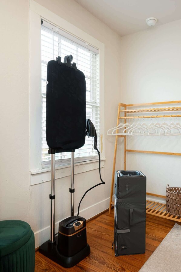 Una habitación con ventana, un aspirador, un tendedero y un otoman verde.