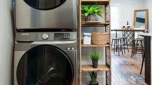 Uma lavanderia moderna com duas máquinas de lavar e secar de aço inoxidável, uma prateleira de vime com objetos decorativos, e uma área de refeições ao fundo.