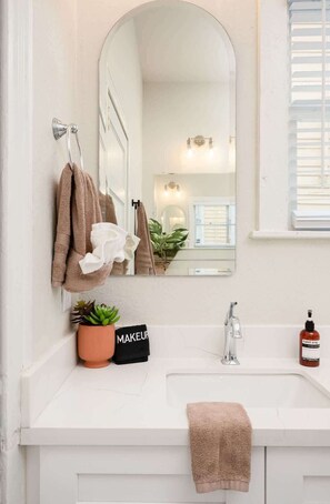 A bathroom with a white sink, a mirror, and a window with blinds. There are towels hanging on a rack and a potted plant on the counter. - Ultimate 2x Home Retreat – Hot Tub, Sauna, Games (Galveston)
