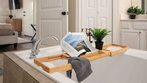 A bathroom with a white bathtub, a wooden tray holding a magazine, a towel, and a potted plant. - Modern Entertainer's Dream: Pool | Cabana | Games (Humble)