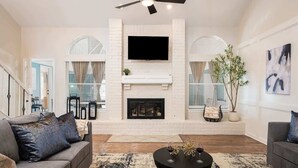 A spacious living room with a fireplace, a ceiling fan, and a large TV mounted above it. - Modern Entertainer's Dream: Pool | Cabana | Games (Humble)