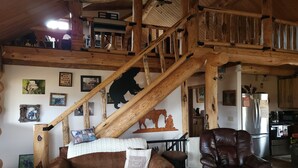 A rustic interior with a wooden staircase, a sofa, a leather chair, and various wall decorations including framed pictures and a bear silhouette. - Charming 2-bedroom cabin in Emigrant with Beautiful Views (Emigrant)