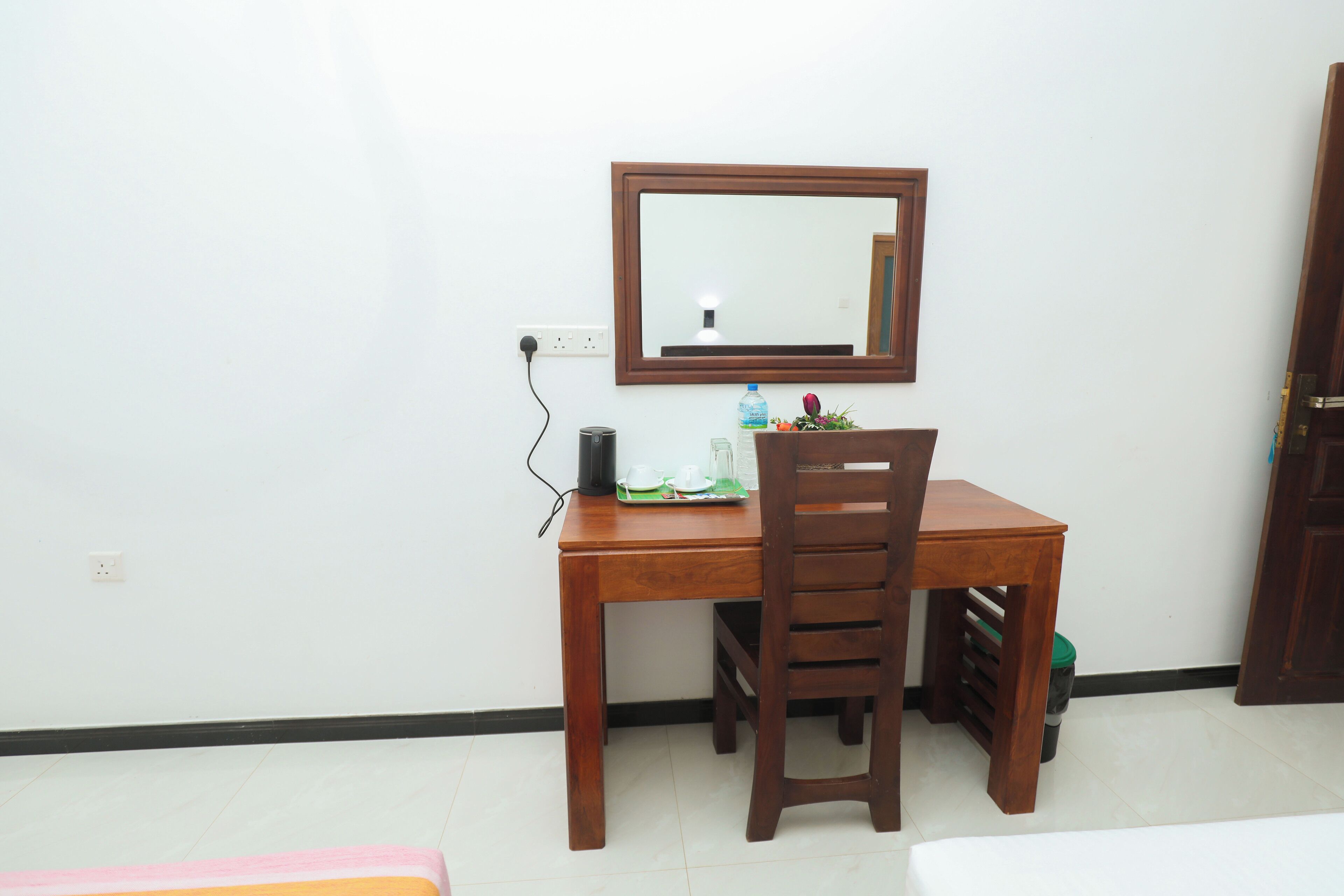 Quarto pequeno e bem organizado, com uma escrivaninha de madeira, uma cadeira, um espelho e uma lixeira.