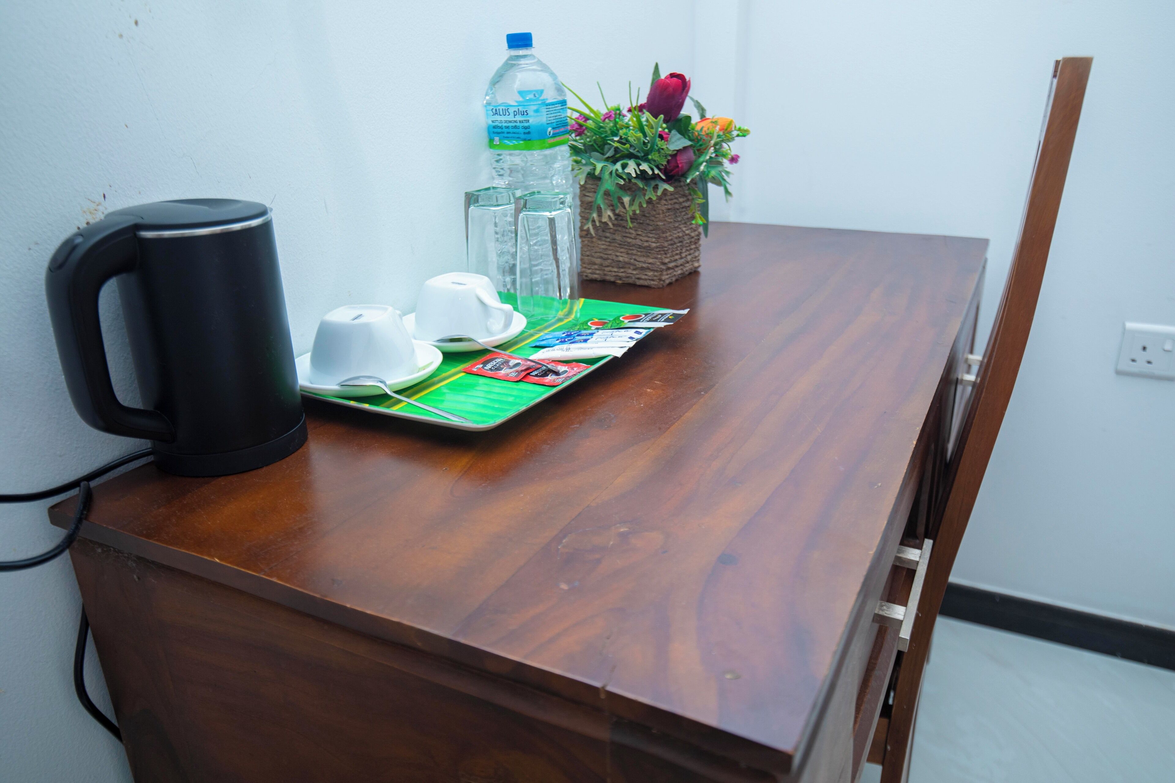 Una mesa de madera con un hervidor eléctrico negro, dos tazas blancas, una botella de agua y un juego de cartas.