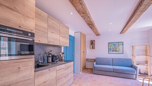 A modern kitchen with wooden cabinets, a built-in oven, and a blue sofa in a living area with a tiled floor. - IN THE HEART OF THE TOWN CENTRE – STUDIO – CLOSE TO THE BEACH (STE MAXIME)