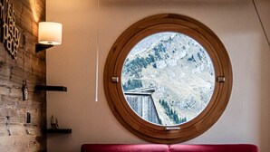 A cozy room with a red sofa, a round window with a view of a snowy mountain, and a wooden wall with a lamp.