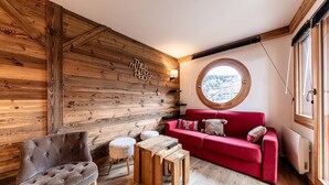 A cozy room with a red sofa, a tufted armchair, and a wooden coffee table. A round window offers a view of a snowy landscape.