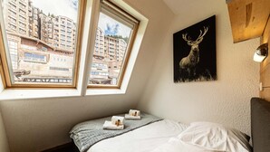Une chambre à coucher avec un lit, deux fenêtres, un tableau représentant un cerf et une vue sur des immeubles.