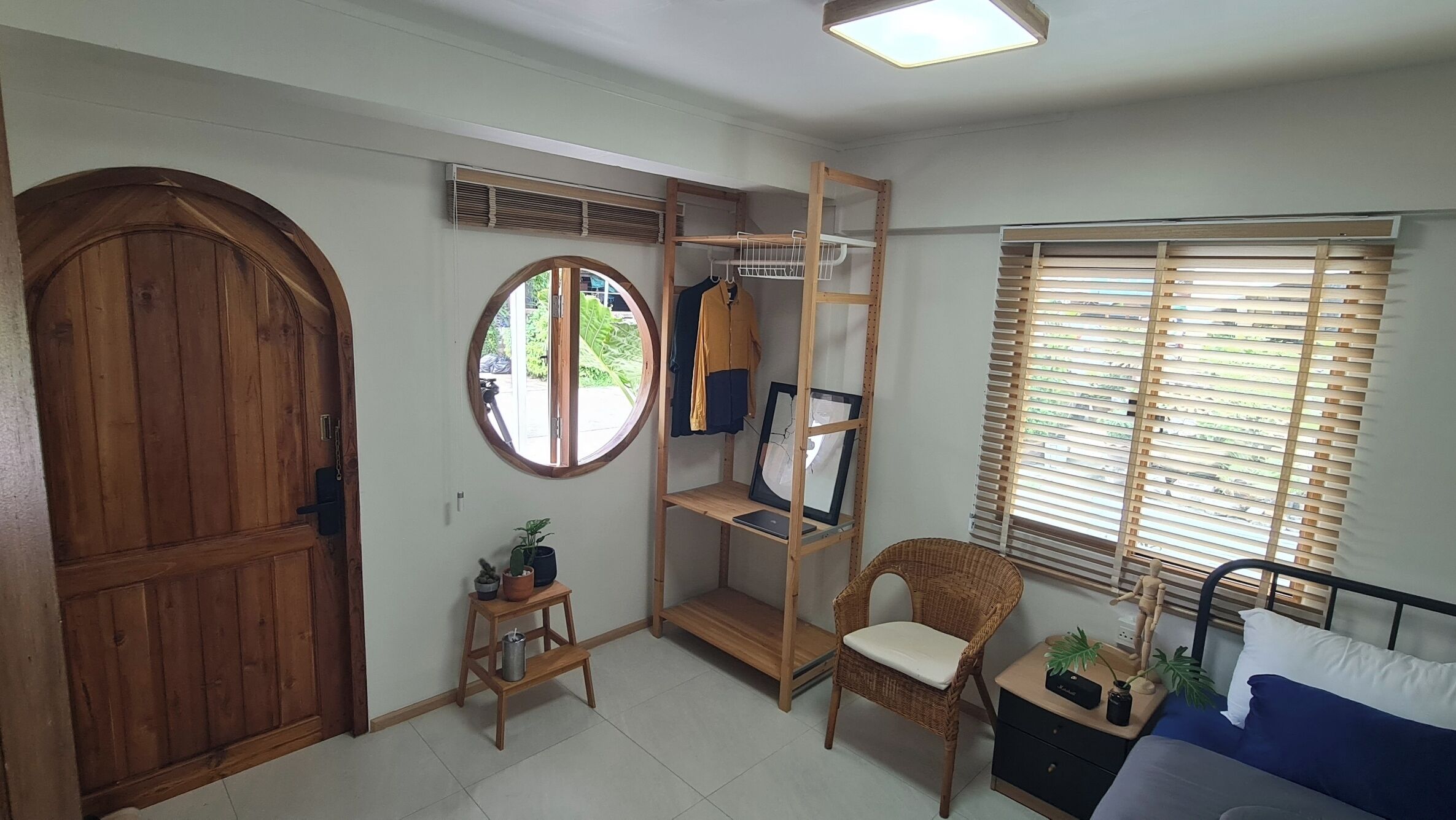 A room with a wooden door, a round window, a built-in shelf with clothes, a chair, a bed, and a window with blinds.