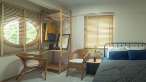 Une chambre à coucher comprenant un lit, deux fauteuils en osier, une table de chevet, une étagère en bois avec des vêtements et une fenêtre avec des stores.