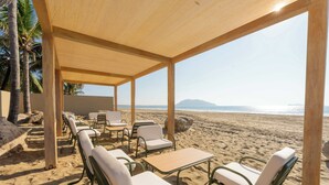 A beachside seating area with tables and chairs under a wooden canopy, overlooking the beach and ocean.