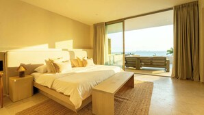A spacious bedroom with a large bed, a nightstand, a bench, and a view of the sea through large windows.
