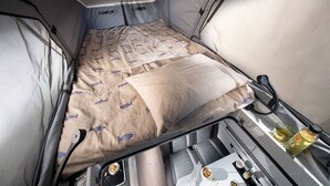 A compact RV interior with a bed, a small table with a bottle and a cup, and a window.