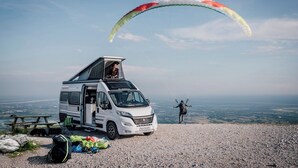 A white van with a rooftop tent and a person preparing to paraglide.