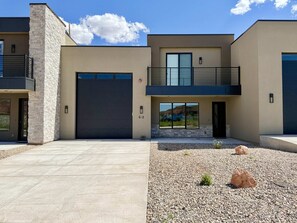 Une maison moderne à deux étages, dotée d’un grand portail de garage, d’un balcon et d’un jardin paysager à l’avant, agrémenté de rochers et de petites plantes.