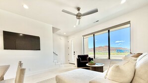 A modern living room with a large window, a ceiling fan, and a flat-screen TV mounted on the wall. - Stylish 3BR Oasis | Pools, Stargazing + Zion/Bryce (Kanab)