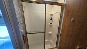 A bathroom with a shower enclosure, a wooden door, and a small shelf with a bottle. - Cozy Camper North Topsail Beach (North Topsail Beach)