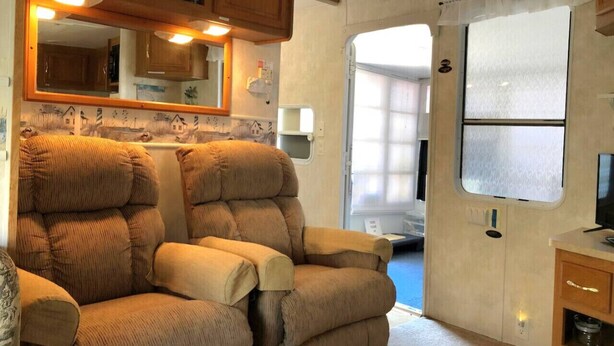 A compact RV interior with a cushioned seating area, a small kitchenette, and a window providing natural light. - Cozy Camper North Topsail Beach (North Topsail Beach)