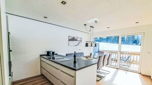 A modern kitchen with a central island, wooden flooring, and a view of a snowy landscape through large sliding doors. - Alpine Qualitytime by Interhome (Wildschönau)