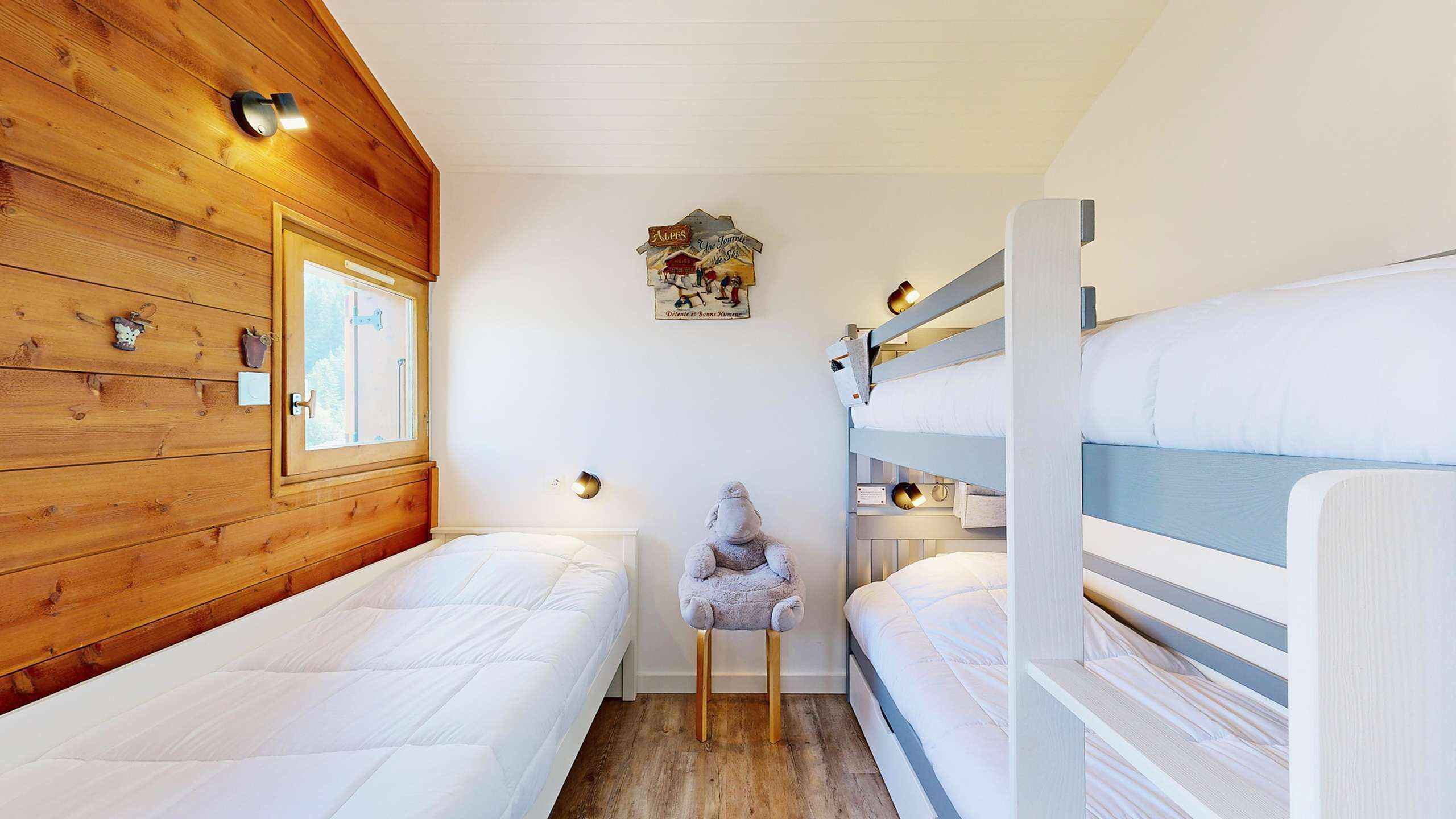 Een gezellig houten kamer, ingericht als blokhut, met een raam, stapelbedden en een klein tafeltje met een lamp.