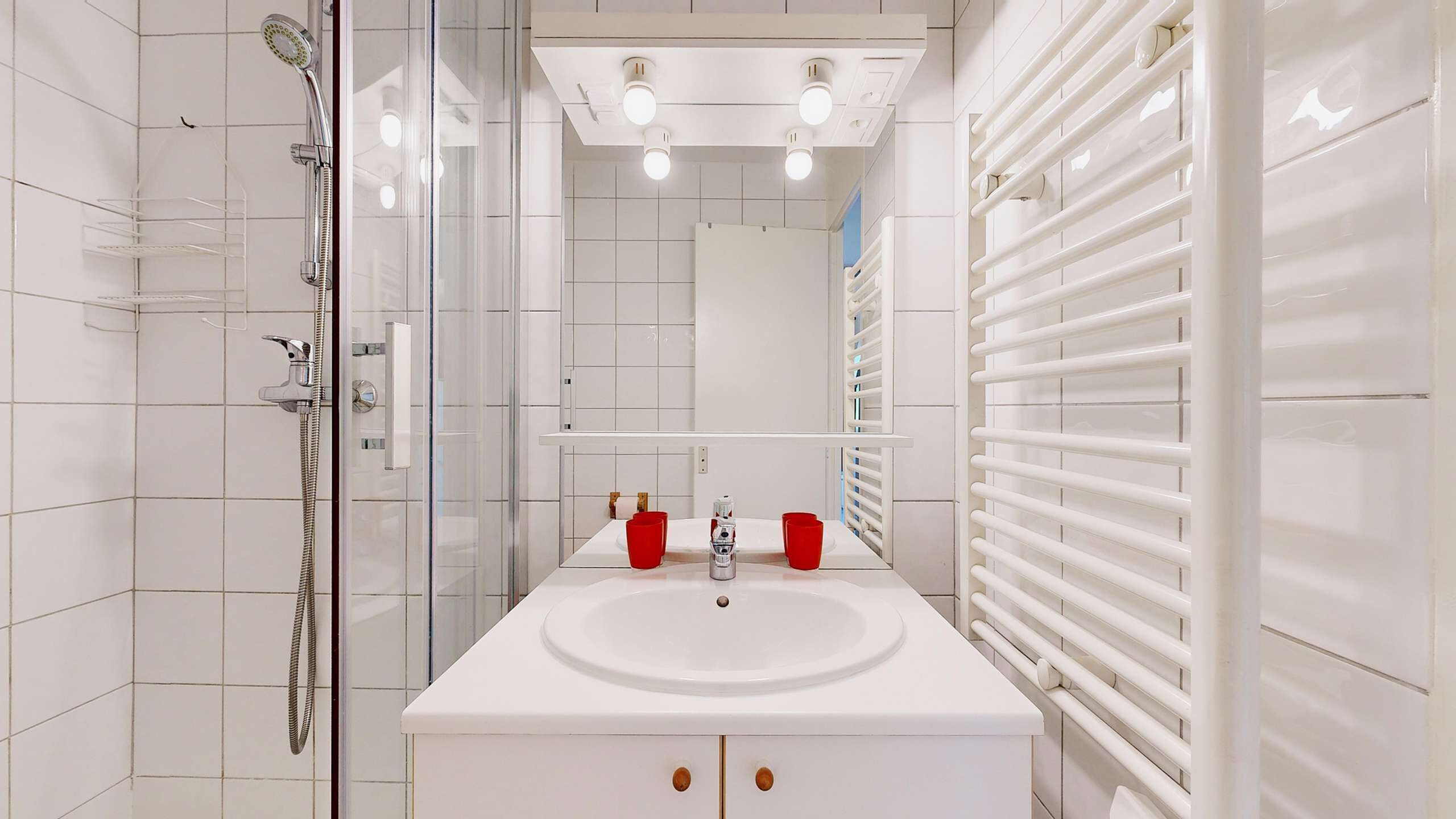 Casa de banho moderna com cabine de chuveiro em vidro, uma pia branca com duas chávenas vermelhas e um porta-toalhas branco.