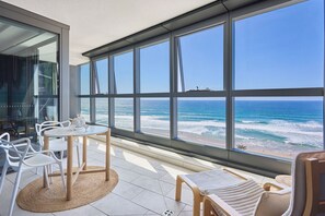 A modern room with large windows overlooking the ocean, featuring a round table and chairs, and a view of the beach. - Jewel Realty (SURFERS PARADISE)