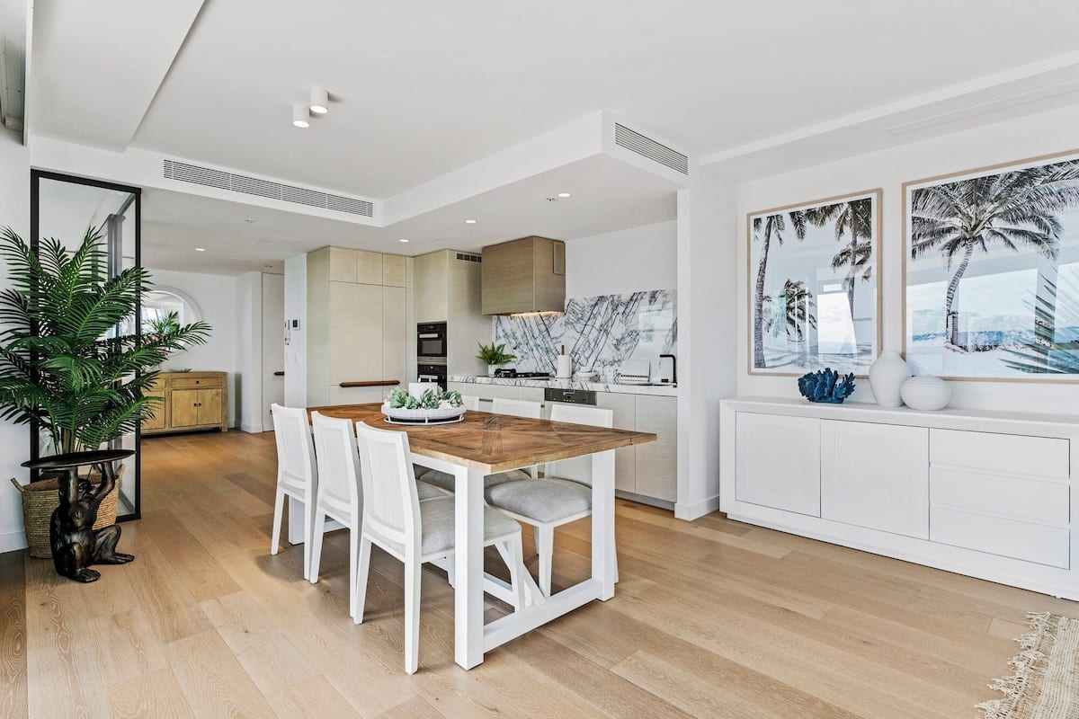 Una cocina moderna con armarios blancos, una mesa de comedor de madera y un amplio ventanal con vista a palmeras.