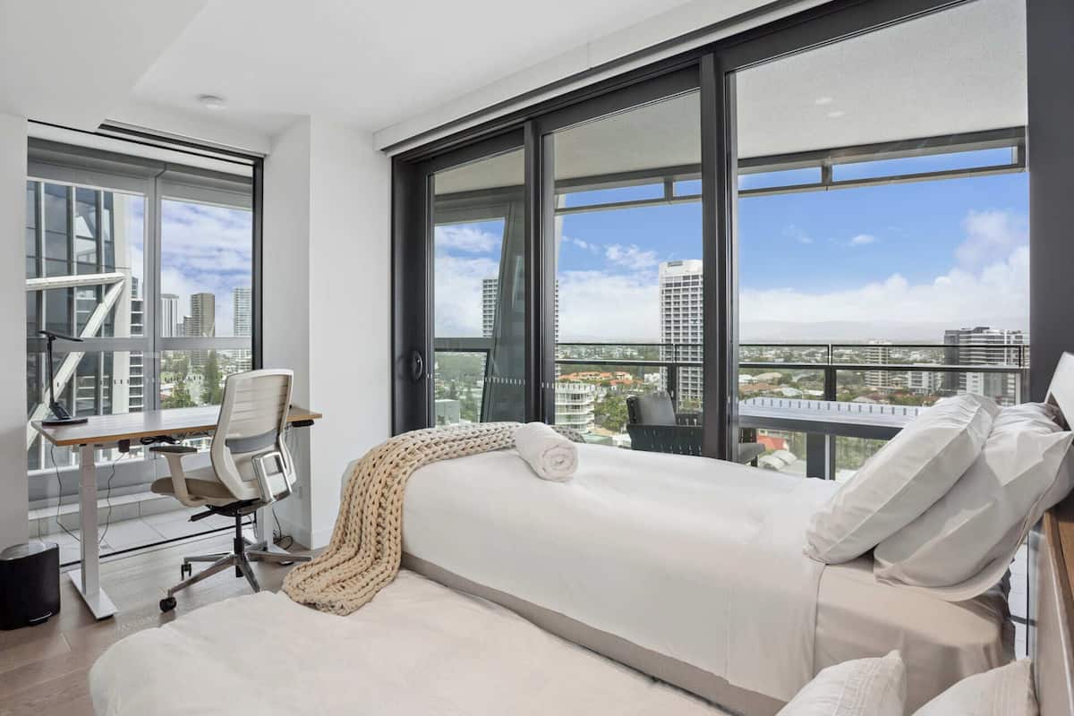 Un dormitorio moderno con una cama grande, un escritorio y una silla, con vistas a la ciudad a través de amplias ventanas.