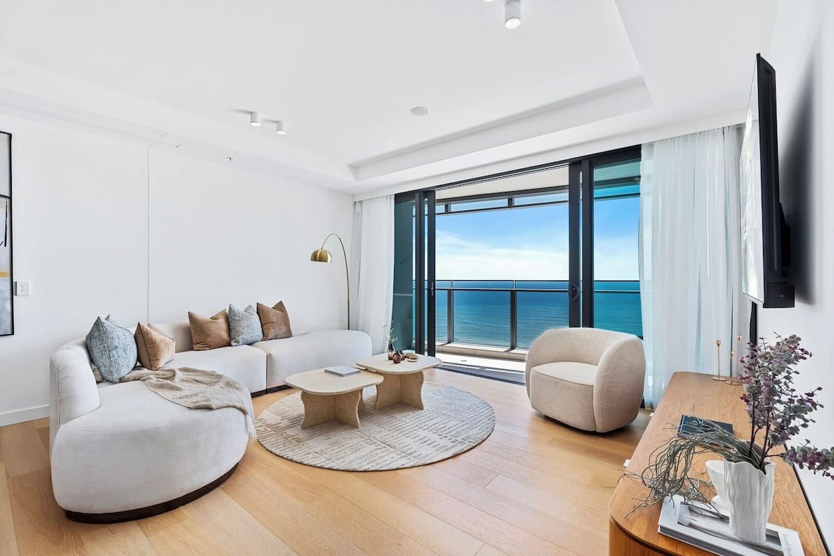 Una sala de estar moderna con un sofá, dos mesas de centro y una silla, un ventanal con vista al mar y un televisor de pantalla plana montado en la pared.