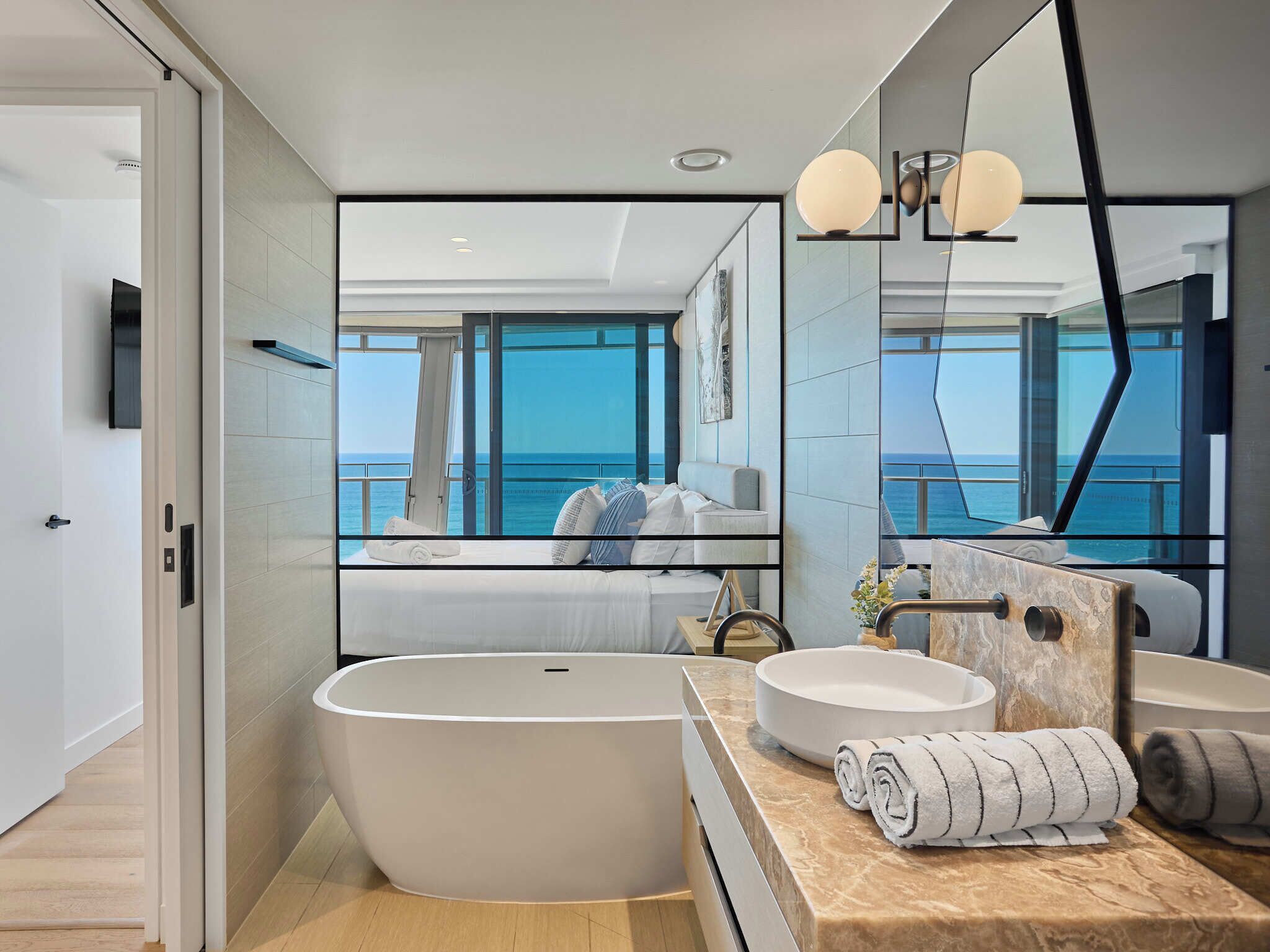 A modern bathroom with a large bathtub, two sinks, and a large mirror reflecting a room with a bed and a view of the sea.