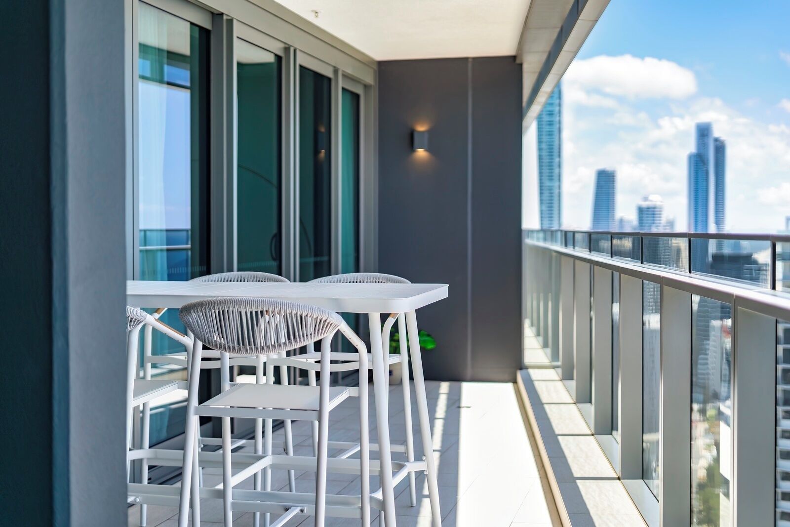 A balcony with white chairs and a table, overlooking a cityscape with tall buildings.