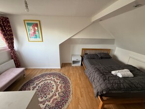 Makuuhuone, jossa on sänky, yöpöytä, penkki, patteri, ikkuna, jossa on verhot, ja seinällä oleva kehystetty kuva.