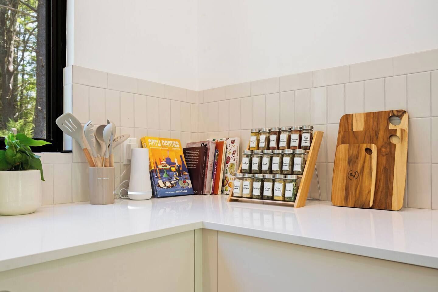 Un plan de travail de cuisine avec un pare-feu en carreaux blancs, une plante en pot, un appareil de cuisine blanc, un livre intitulé « ARTY PARTIES », un porte-épices et une planche à découper en bois.