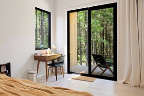 A bedroom with a bed, a desk, and a chair, a large window with a view of a forest, and a sliding glass door leading to a balcony with a chair.