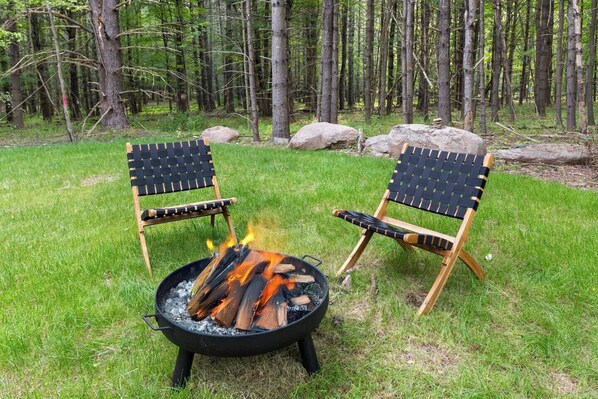 Two Adirondack chairs facing a campfire in a grassy area with trees in the background.