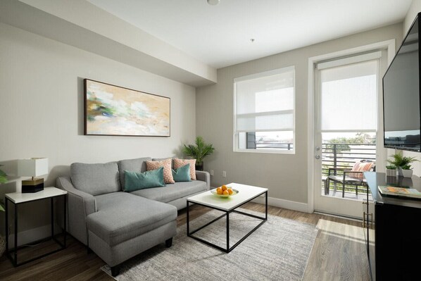 A modern living room with a grey sofa, a coffee table, a painting on the wall, and a balcony with a view. - The Mandarin @ Main Place - Free Disney Gift Card! (Orange)