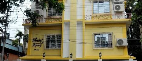 Un edificio de dos pisos con fachada amarilla y blanca, múltiples ventanas y unidades de aire acondicionado.