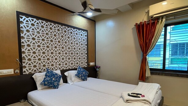 A hotel room with two beds, a decorative wall panel, a ceiling fan, a window with curtains, and a view of buildings outside. - Green Leaf Regency  (Kolkata)