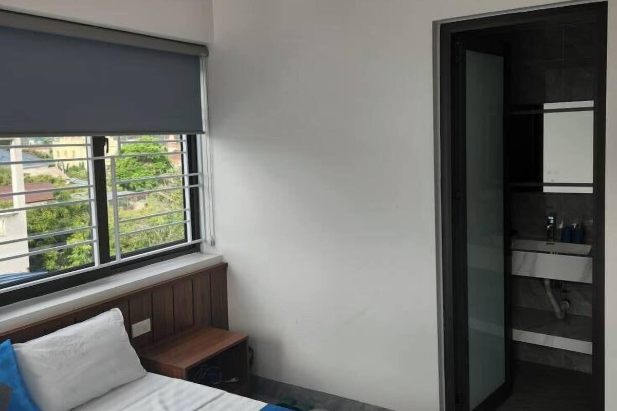 Habitación de hotel con cama, ventana con persianas, equipo de aire acondicionado y baño visible a través de una puerta abierta.