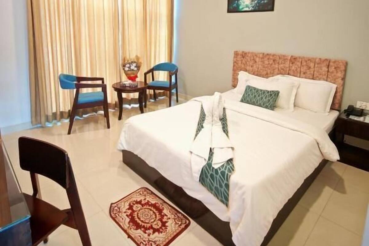 Une chambre d’hôtel avec un lit, deux chaises, une table, un ventilateur de plafond, un tableau et un tapis à motifs.