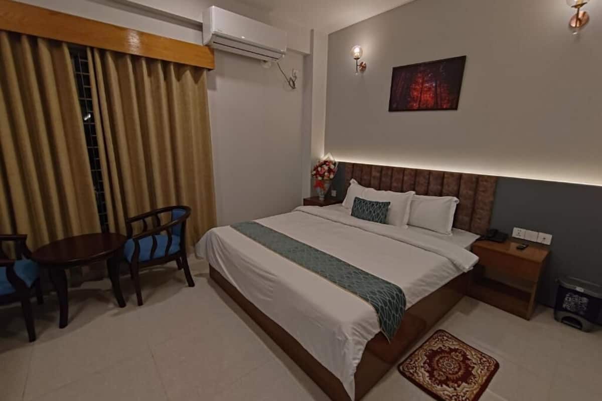 Une chambre d’hôtel comprenant un lit, un bureau, une chaise, une petite table, une table de chevet, une lampe fixée au mur et un tableau accroché au mur.
